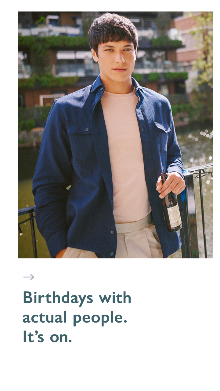 Model in navy shacket and white t shirt at a birthday party holding a bottle of beer standing by a river in summer.