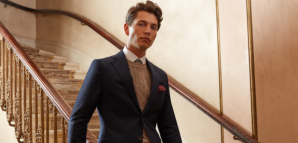 Man dressed in Italian style Moss Bros navy suit with polka-dotted pocket square and a grey patterned jumper.