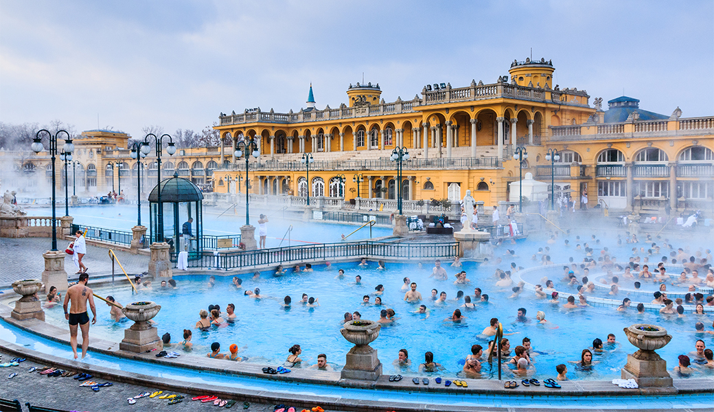 A large crowd of people bathe in Budapest's ancient thermal spa during the summer. There is a grand mustard coloured building in the background.