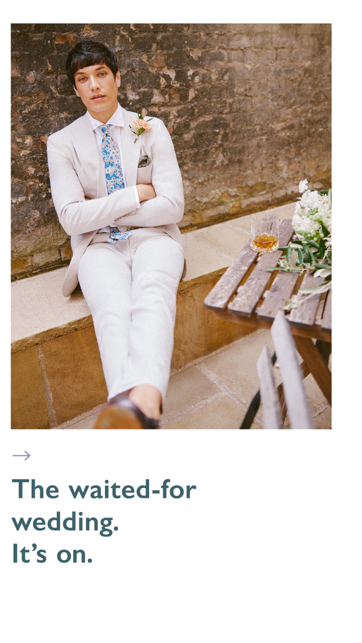 Man in white summer wedding suit and a floral tie with his legs crossed. Text underneath reads 'the waited-for wedding. It's on.'