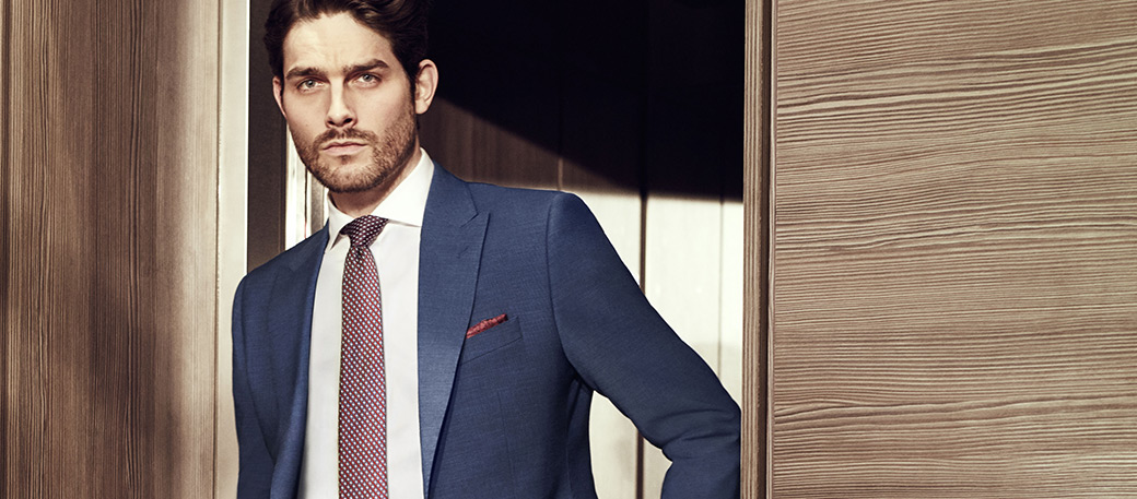 Man with stubble wearing a navy blue Moss Bros 24 hour performance suit with a maroon pocket square and a maroon tie.