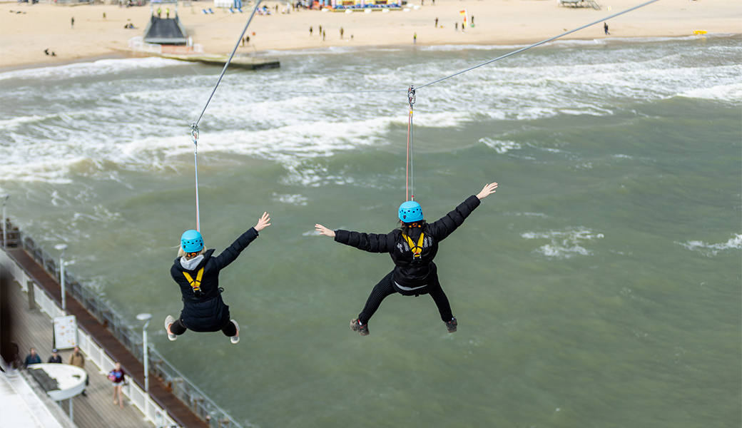 Two men on a summer stag do with arms outstretched ride a zipline in Bournemouth.