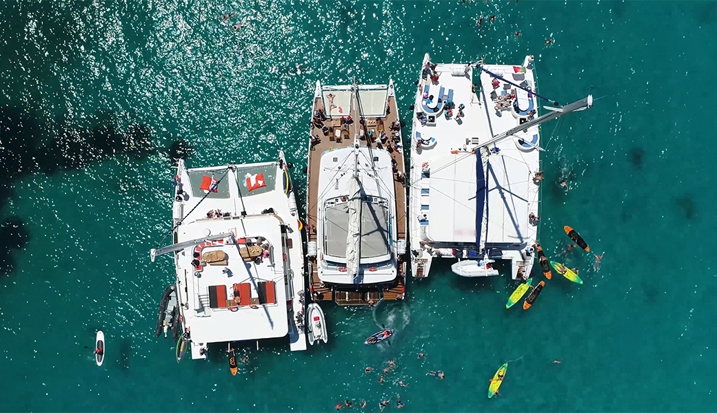 Three catamarans in the sea with kayaks surrounding them, with people on a summer stag do standing on deck.