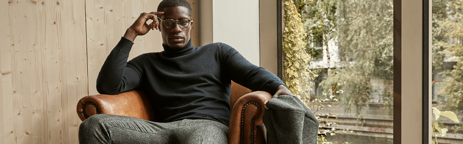 Man in glasses sitting on a leather chair whilst looking at the camera. He is wearing a black merino roll neck jumper and grey tweed trousers.