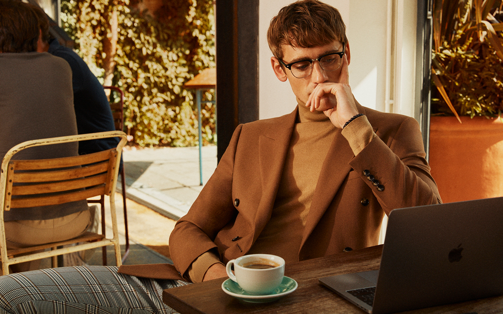 Model sat at his desk in a sunny cafe with a coffee wearing a tan overcoat and tan merino rollneck.