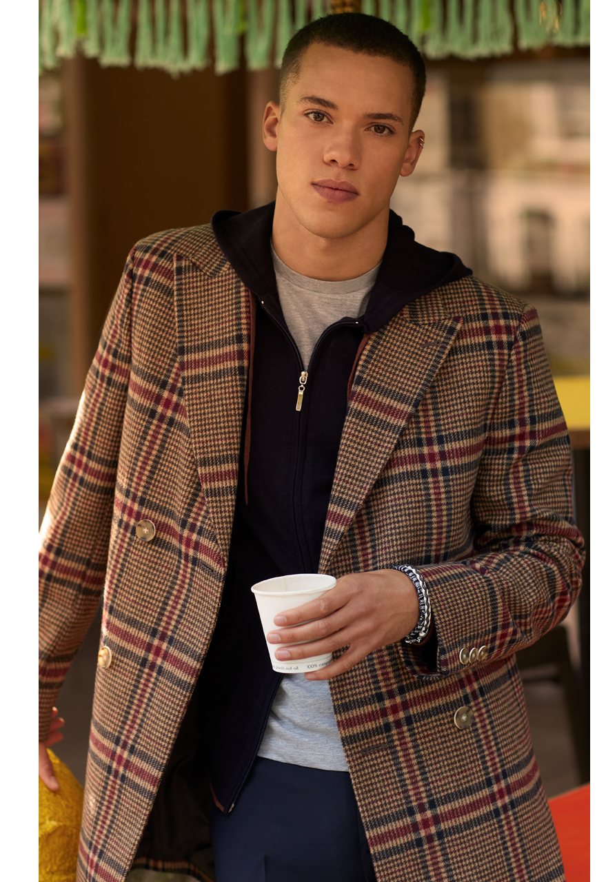 Man holding a coffee cup wearing a Moss Bros knitted hoodie under a slim fit tan and red checked overcoat.