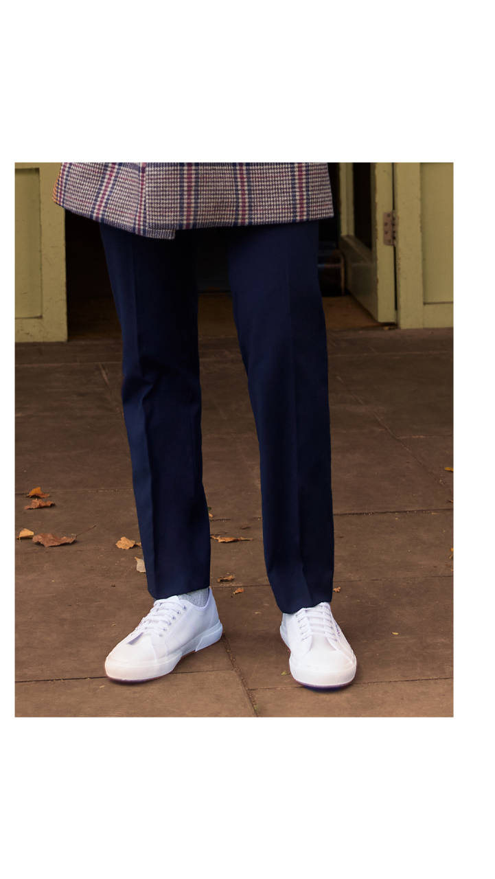 Man wearing Moss Bros slim fit blue stretch trousers , fresh white trainers and a slim fit tan and red checked overcoat.