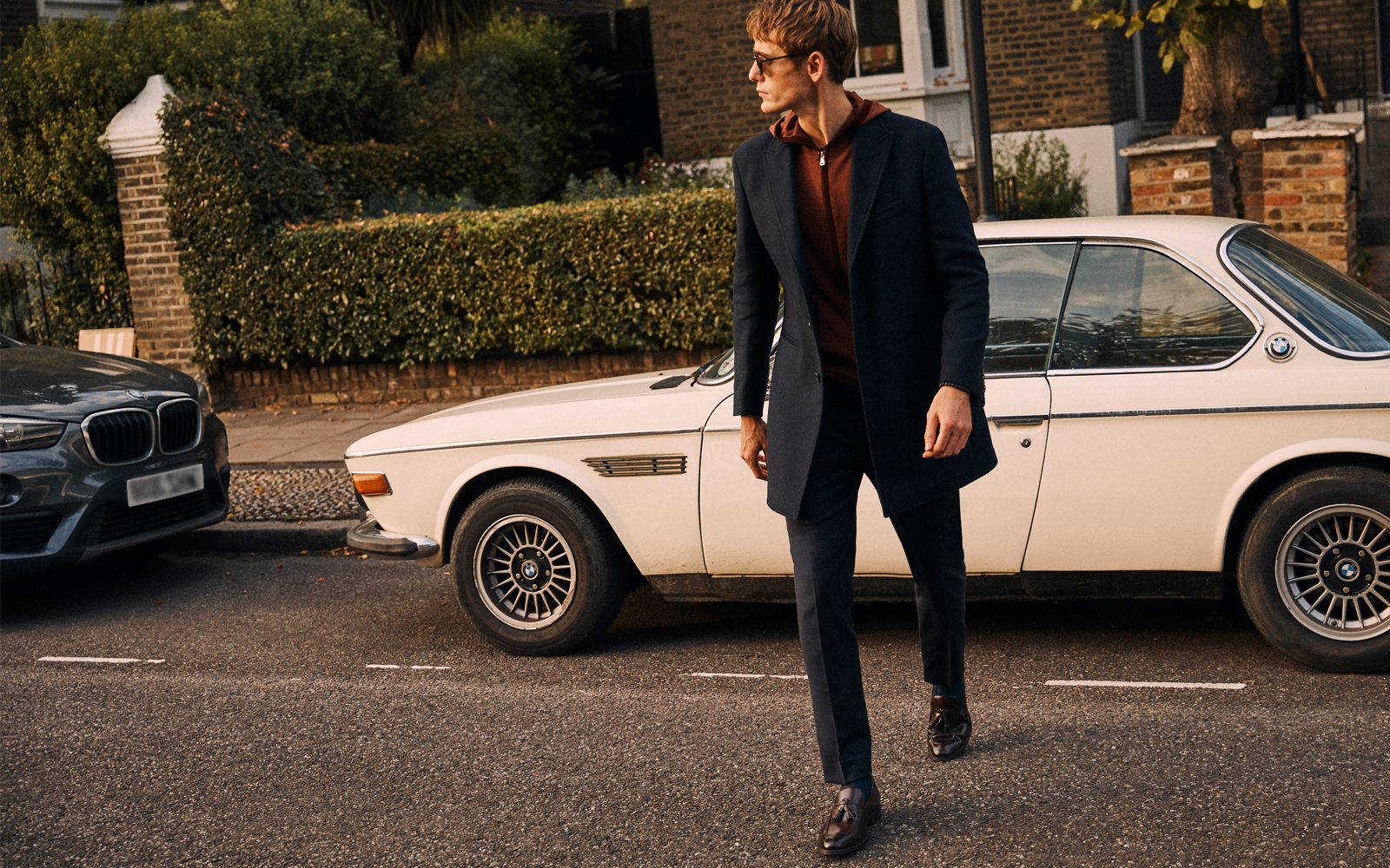 Man crossing the street in front of a classic BMW wearing navy overcoat, smart leather shoes, tailored fit navy brown trousers and a rust Moss Bros merino knitted hoodie.