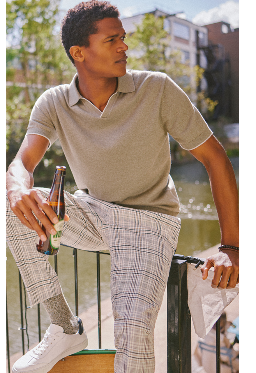 Man in grey merino polo shirt and grey checked trousers leaning on a rail. He is sat by a river and holding a beer in the summer sun.