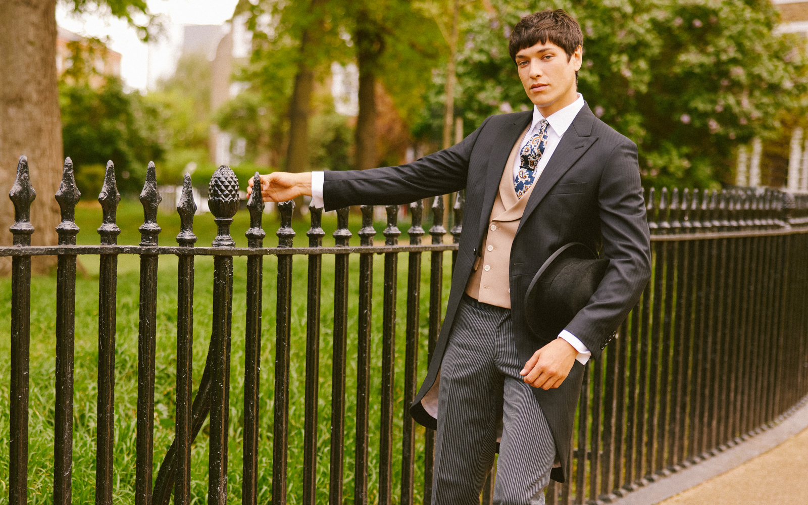 Man in three piece wedding suit and floral tie leaning against park railings in summer.