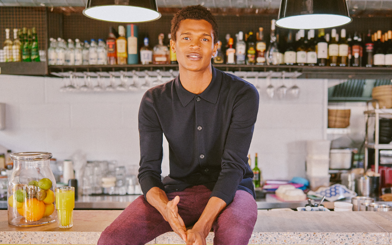 Man sitting on bar top in buttoned black knitted shirt and maroon chinos.