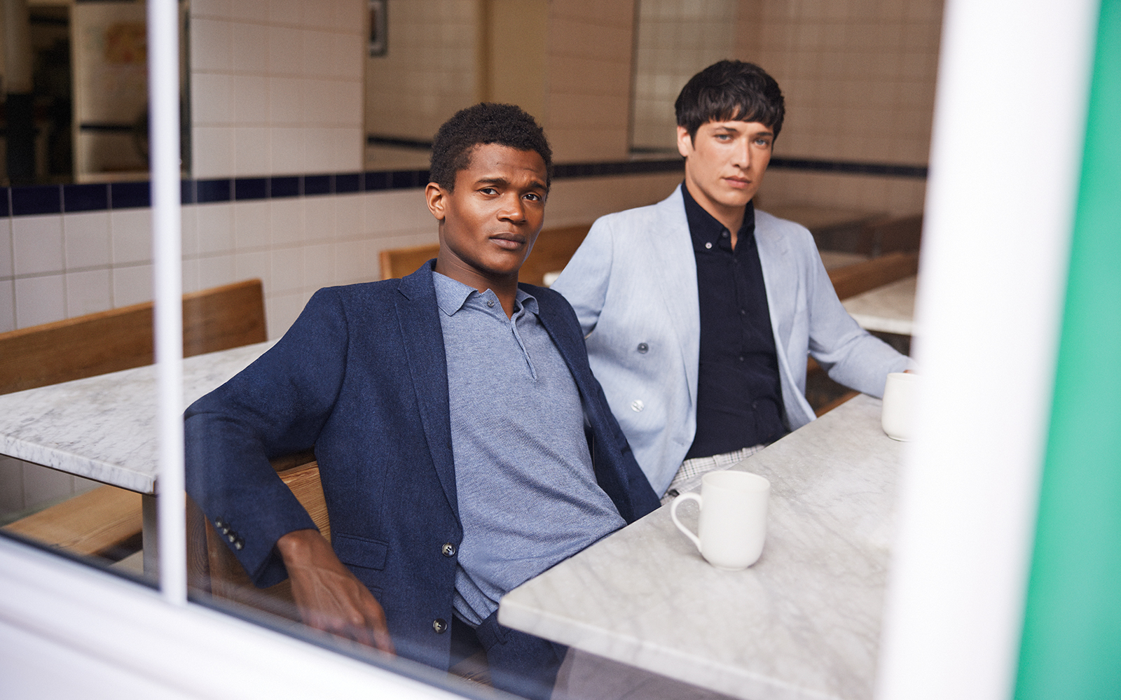 Two men at work; one is wearing a navy blue jacket and grey-blue merino polo shirt and the other is wearing a light grey jacket and dark navy shirt.