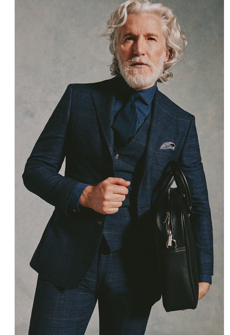 Professional going to work in dark checked three piece suit, a dark denim shirt and tie. Under his left arm he is carrying a black leather hold-bag.