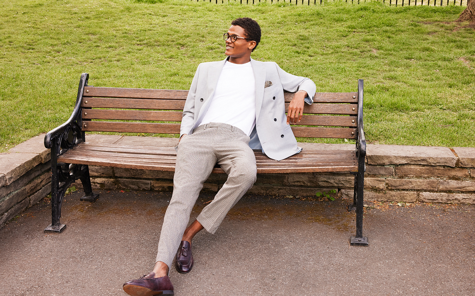 Man at work sitting on a park bench wearing a white t shirt, leather shoes and a light grey work suit.